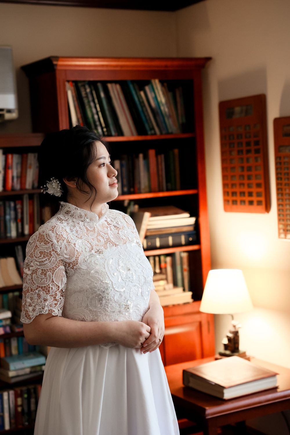 A bride in an elegant white lace dress stands in a library room with wooden bookshelves and warm lighting. smilein wedding photography