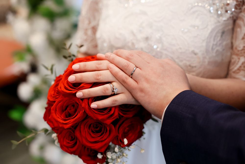 Hands wearing a wedding ring hold a vibrant red rose bouquet against a white lace dress. smilein wedding photography