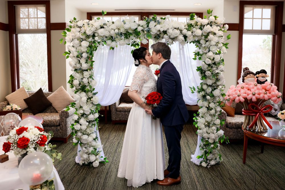 A romantic indoor wedding ceremony under a floral arch decorated with white roses and greenery against a white backdrop. smilein wedding pho