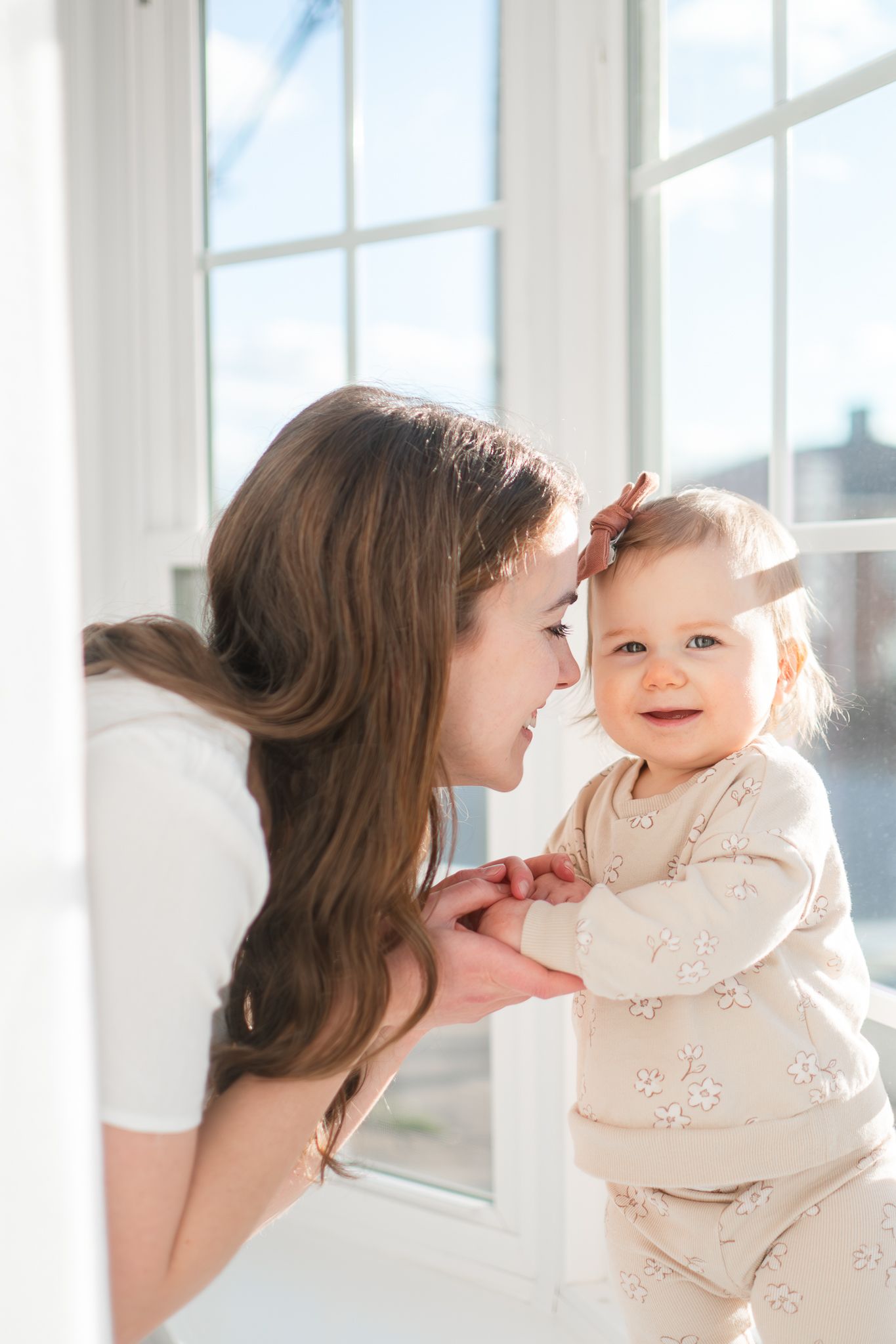 A tender moment between parent and baby by a bright window in matching cream-colored outfits.