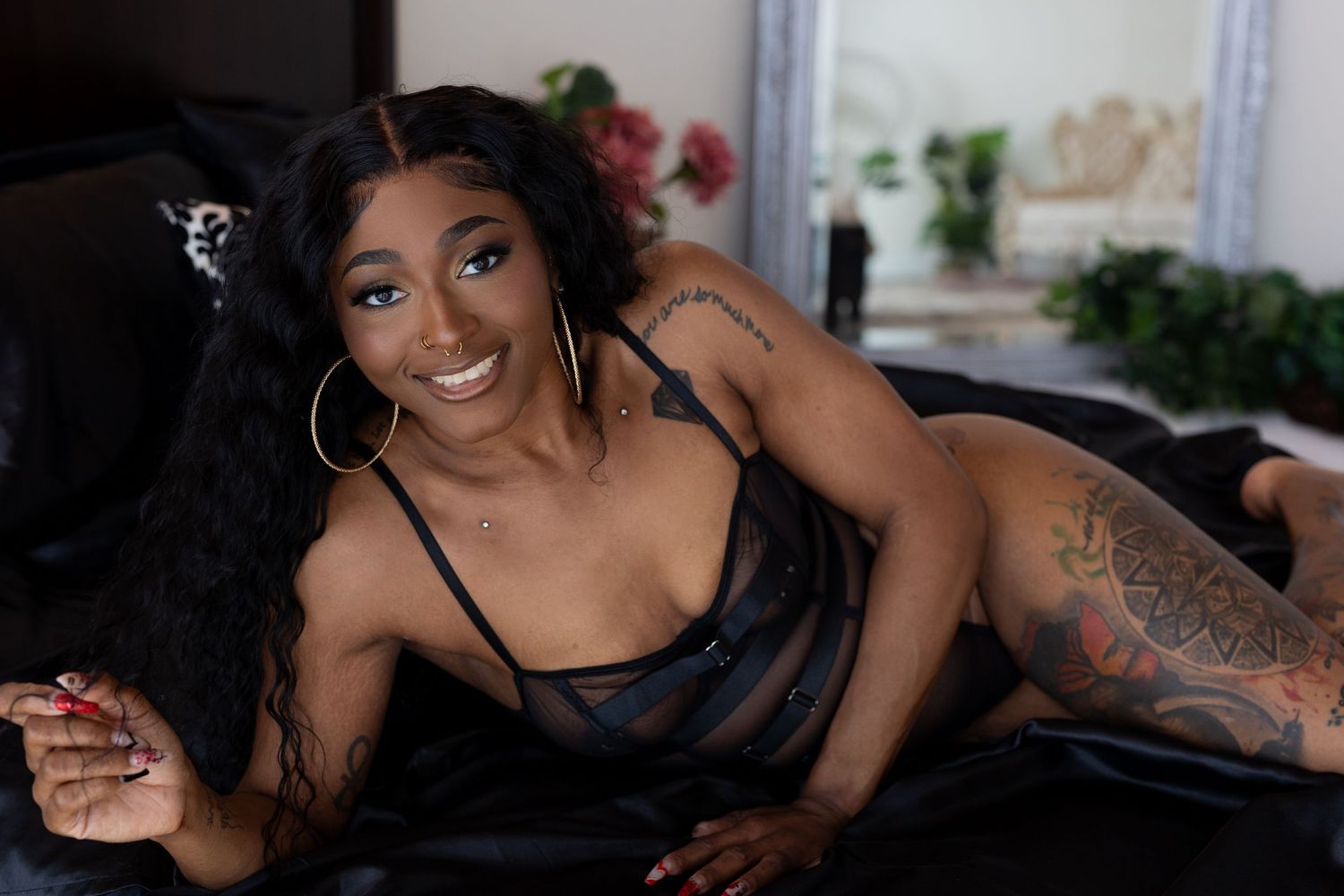 a woman poses seductively on black furniture wearing a dark silk dress and hoop earrings with visible arm tattoos.