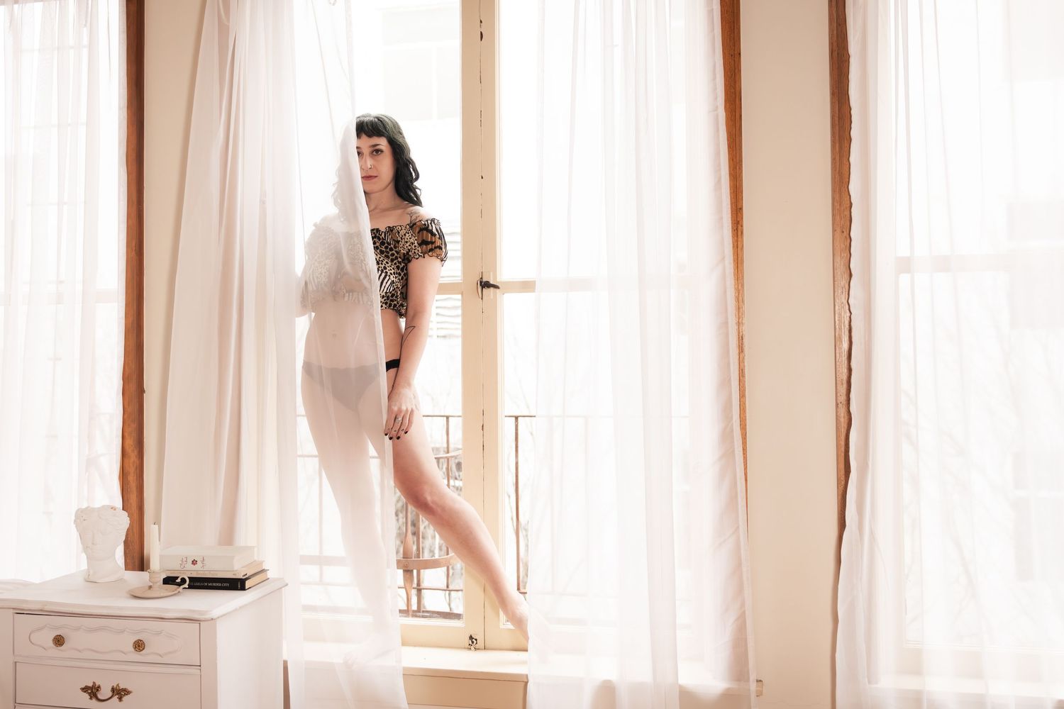 A person in a white robe stands by sheer curtained windows while holding a cat in their arms.