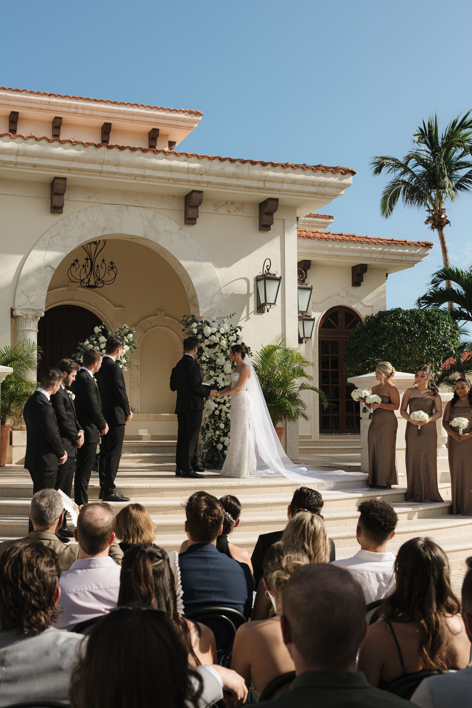 Wedding ceremony at Spanish-style venue with palm trees and elegant architecture.