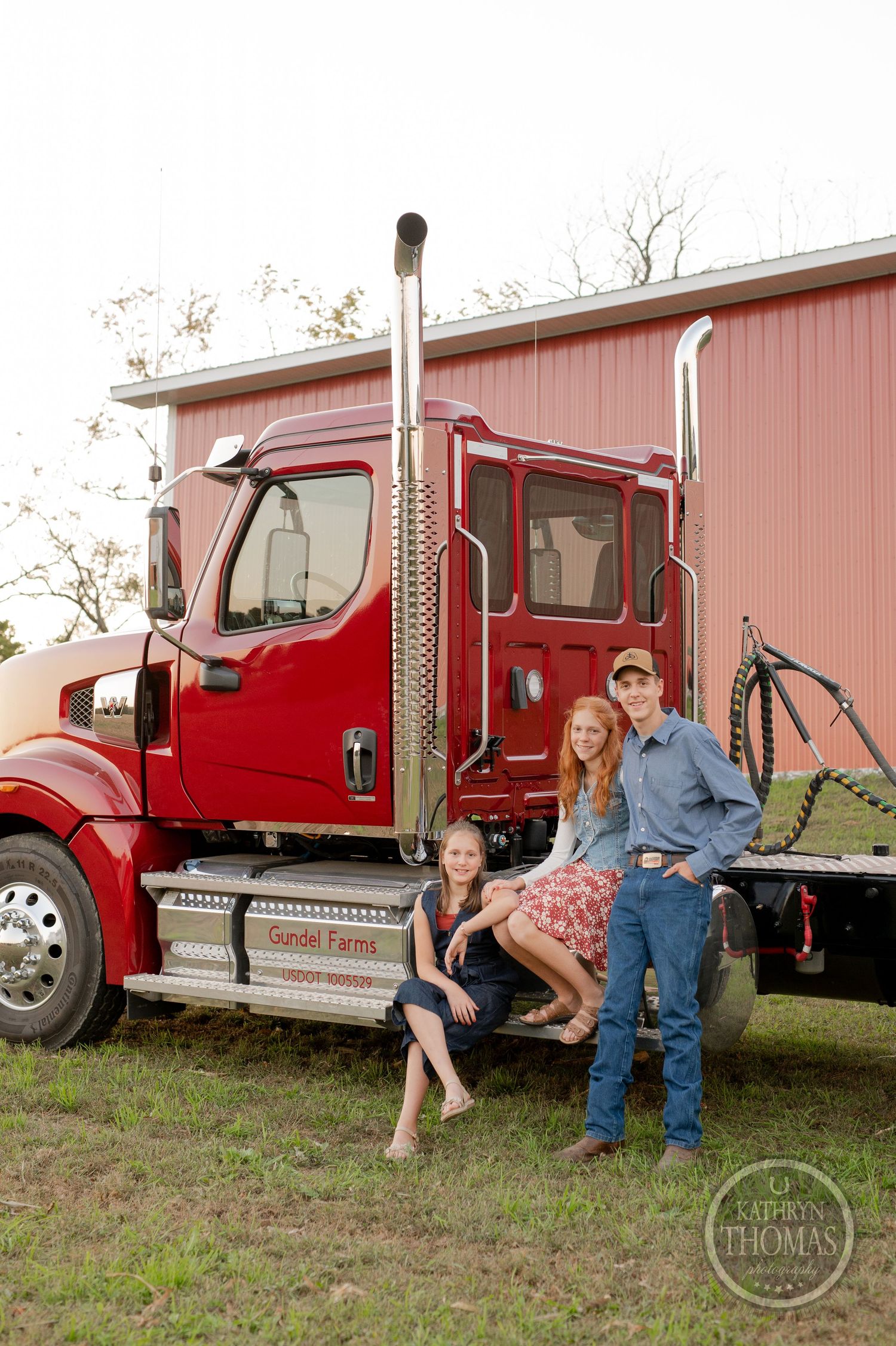 Three people pose together on the steps of a red semi truck parked by a barn.