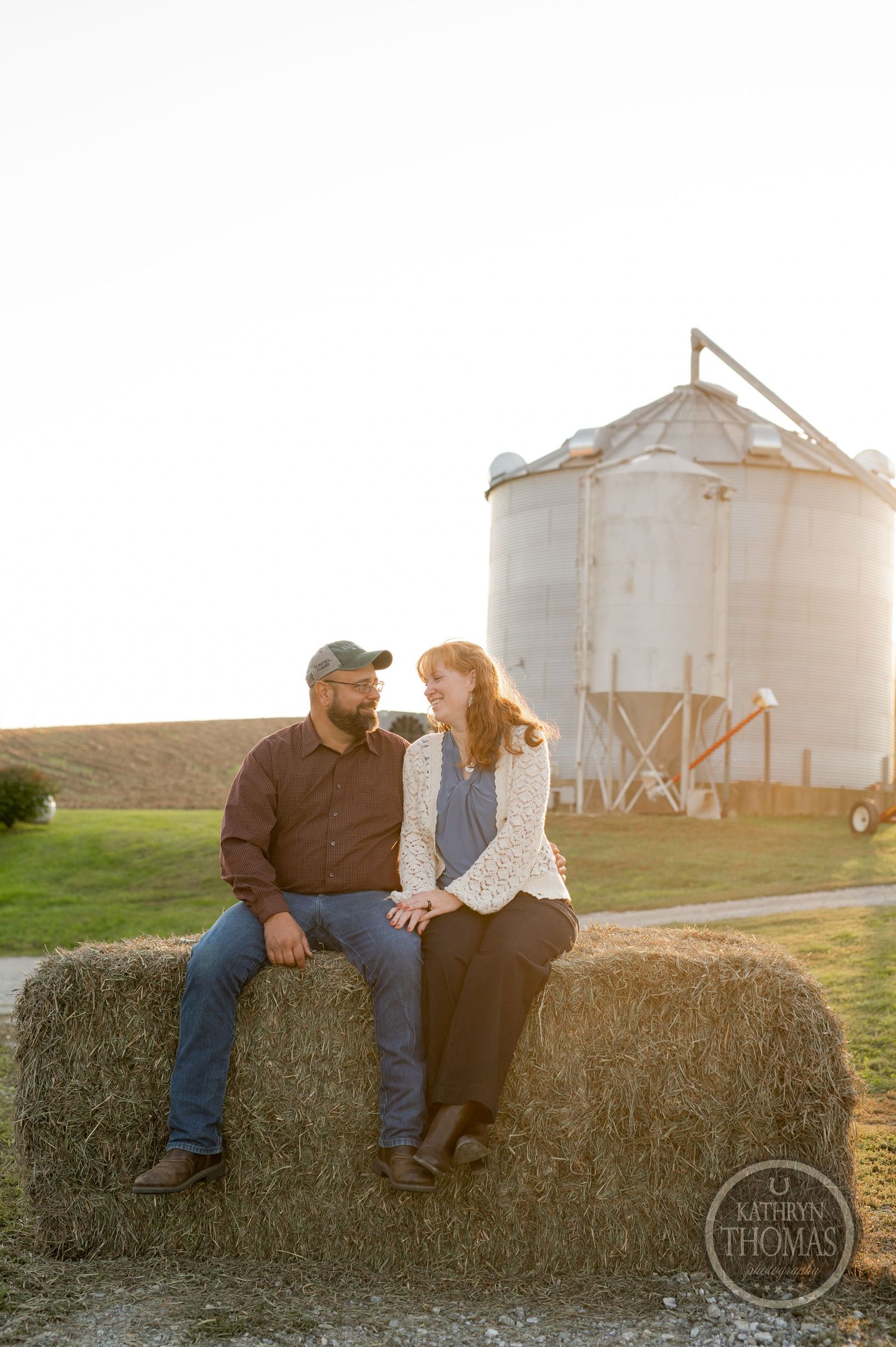 Couple sits together on hay bale with grain silo in background during sunset.
