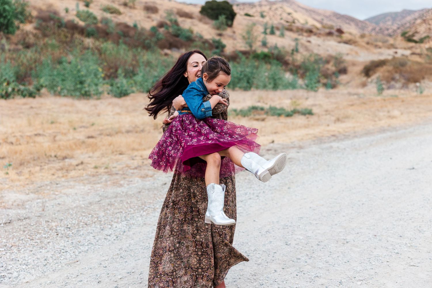 Parent and child in casual clothing having fun and twirling on a rural dirt road against a mountainous backdrop.