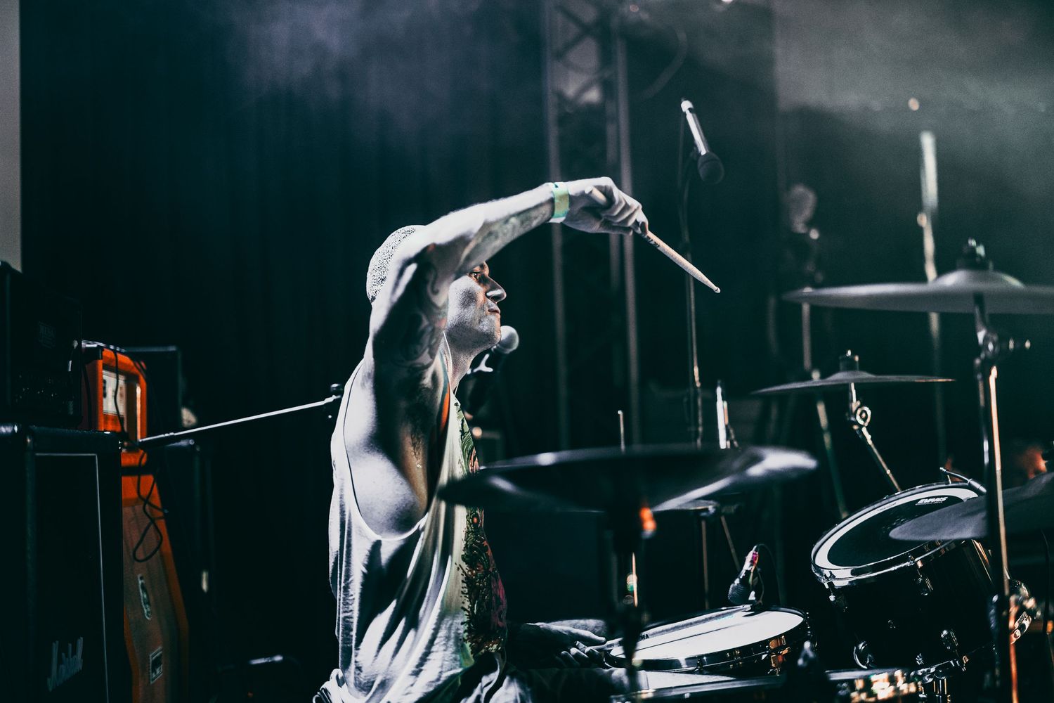 Energetic drummer performs on stage in dramatic lighting with arms raised during an intense musical performance.