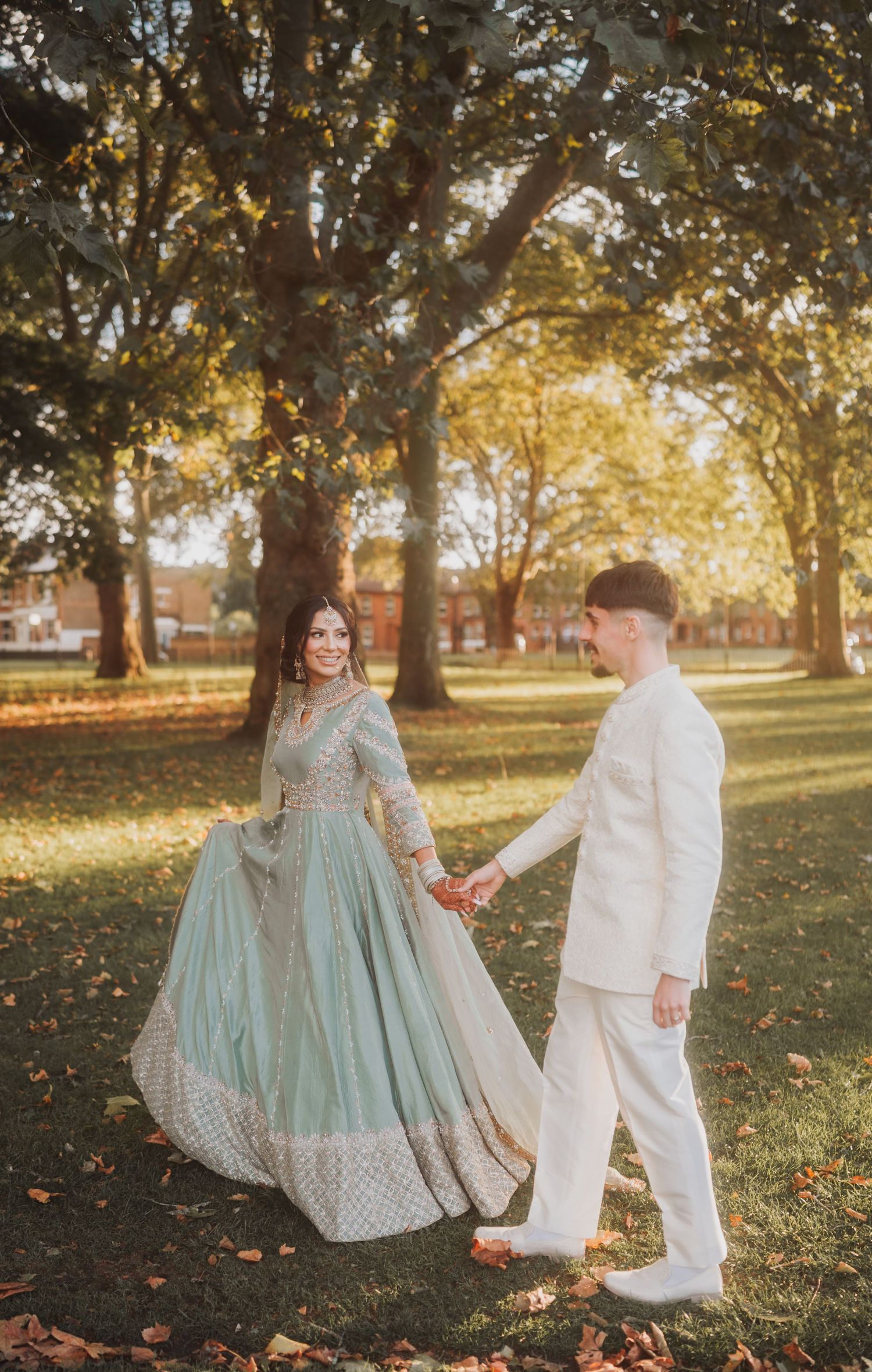 Couple in fusion wedding attire walking through autumn trees in golden sunlight.