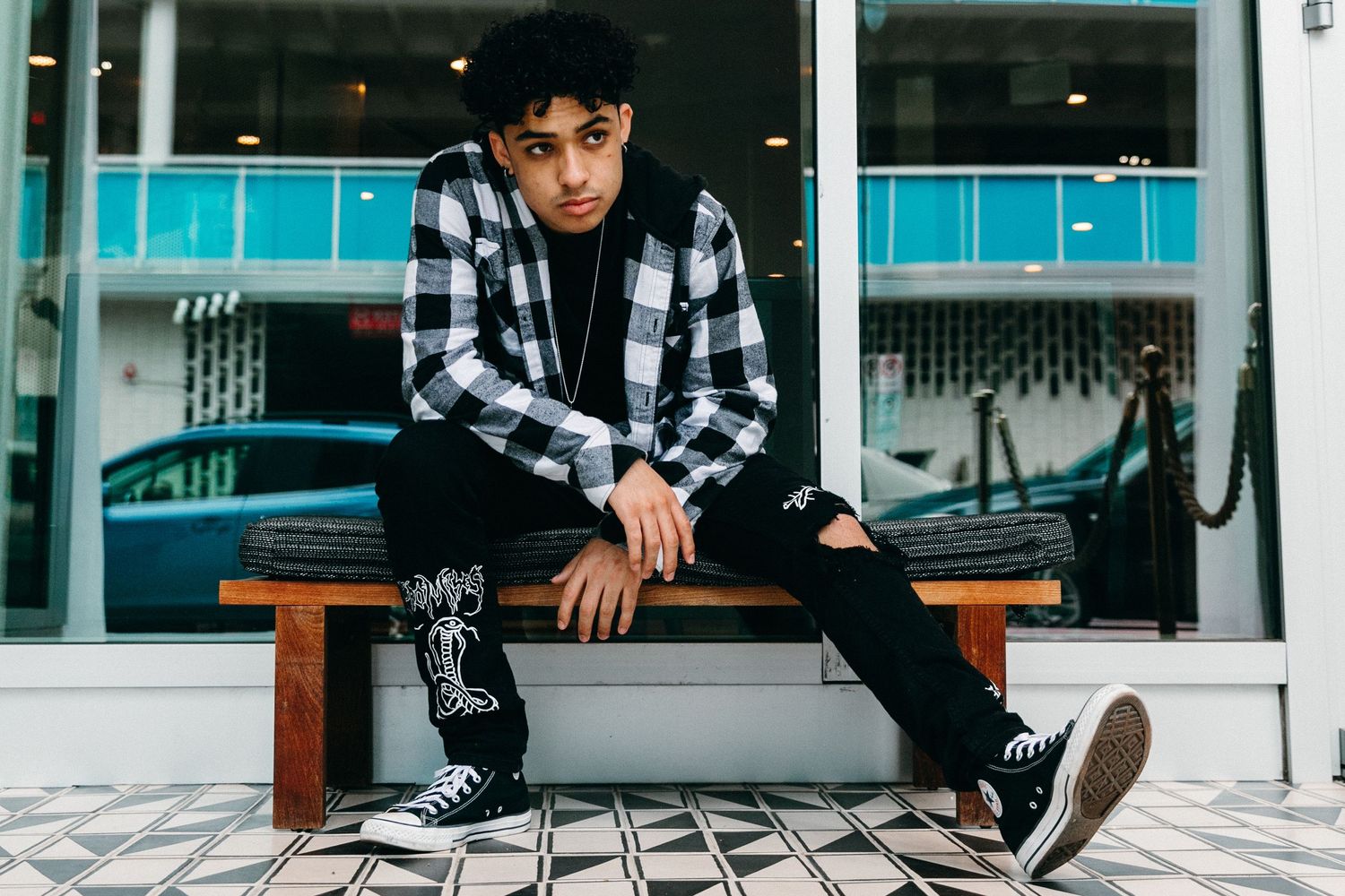 Young person in plaid shirt sitting on wooden bench against modern storefront.