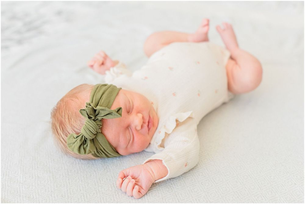 A sleeping newborn wearing a sage green headband and cream colored onesie on white bedding.