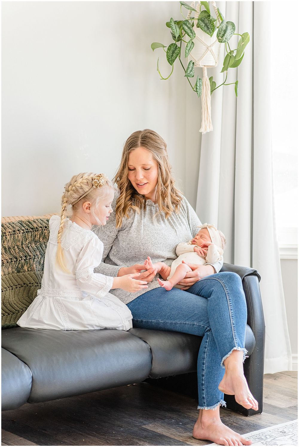 Mother and daughter sharing tender moment with newborn on dark leather couch near hanging plants.