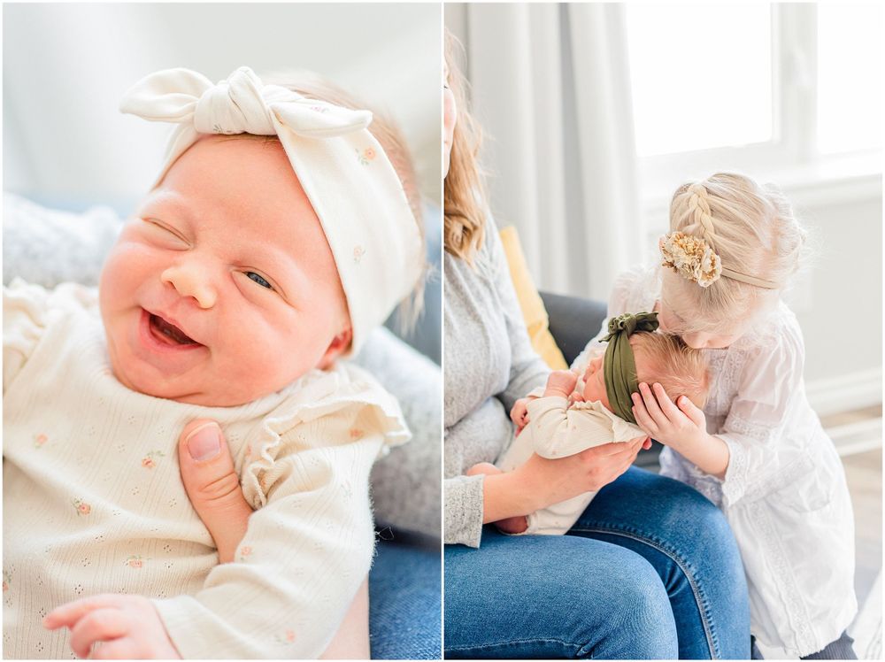 Baby wearing white outfit and bow smiles joyfully while resting on soft surface near window.