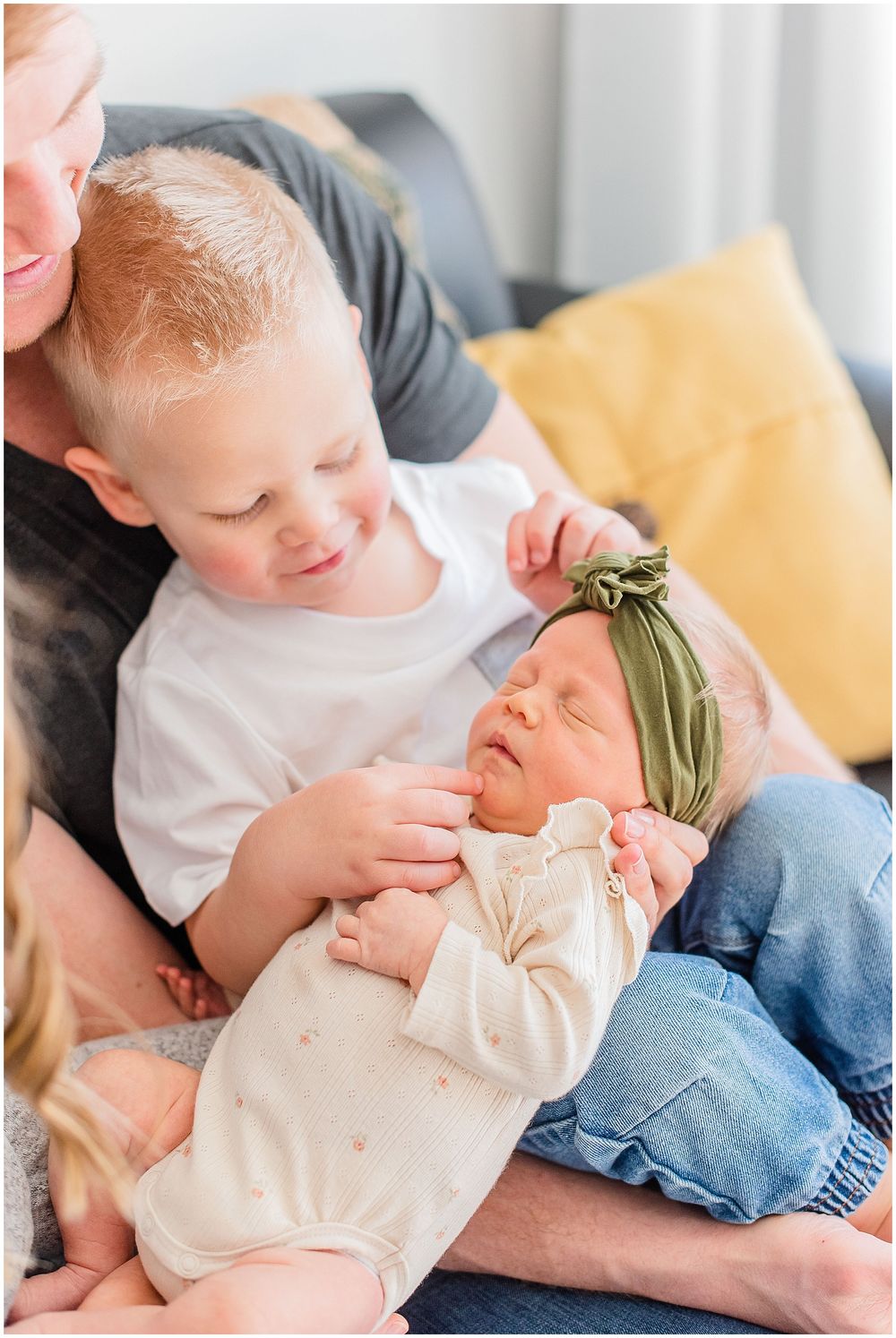 Two siblings share tender moment while sitting on couch as toddler gently touches baby wearing headband.
