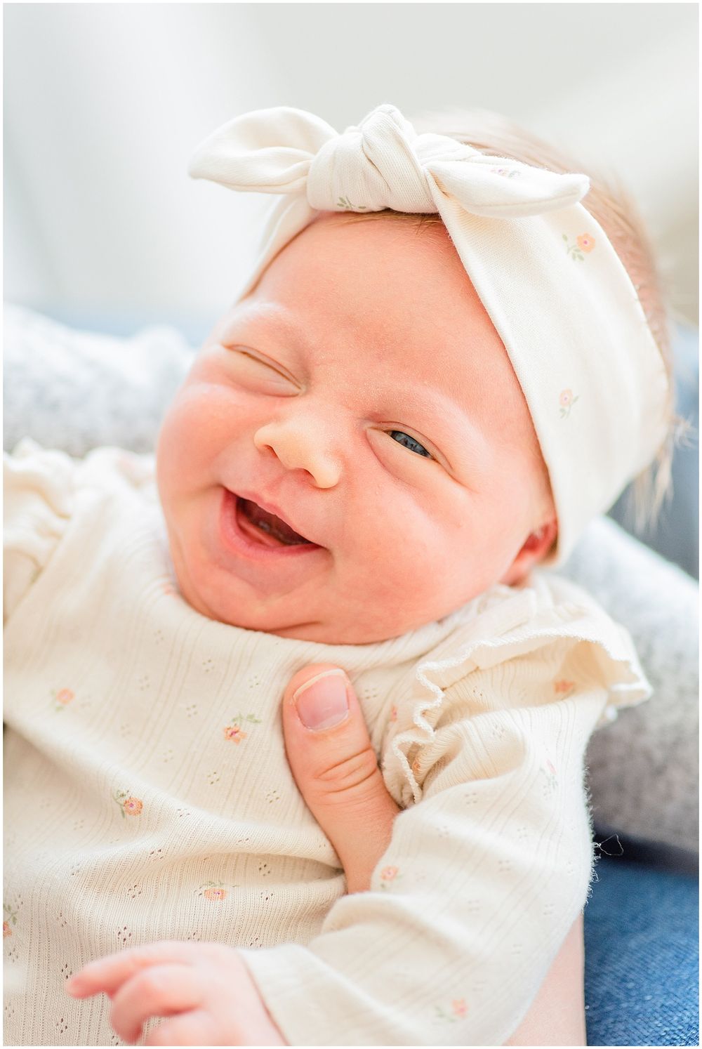 A joyful baby wearing a white bow headband smiles brightly while lying on blue fabric.