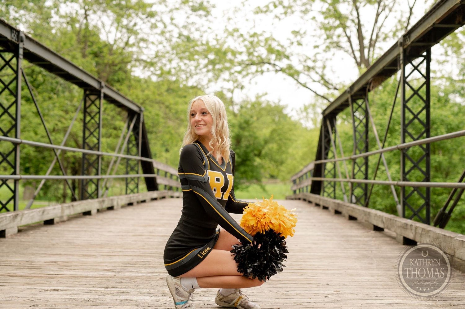 Athletic performer kneels with black and gold pom poms on wooden bridge surrounded by trees.