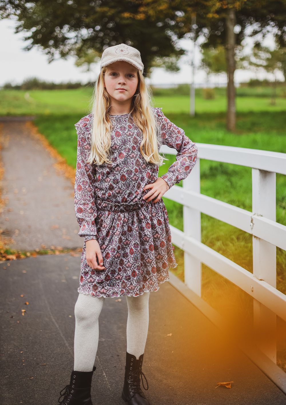 A figure in a floral dress and white tights stands on a path near a white fence.