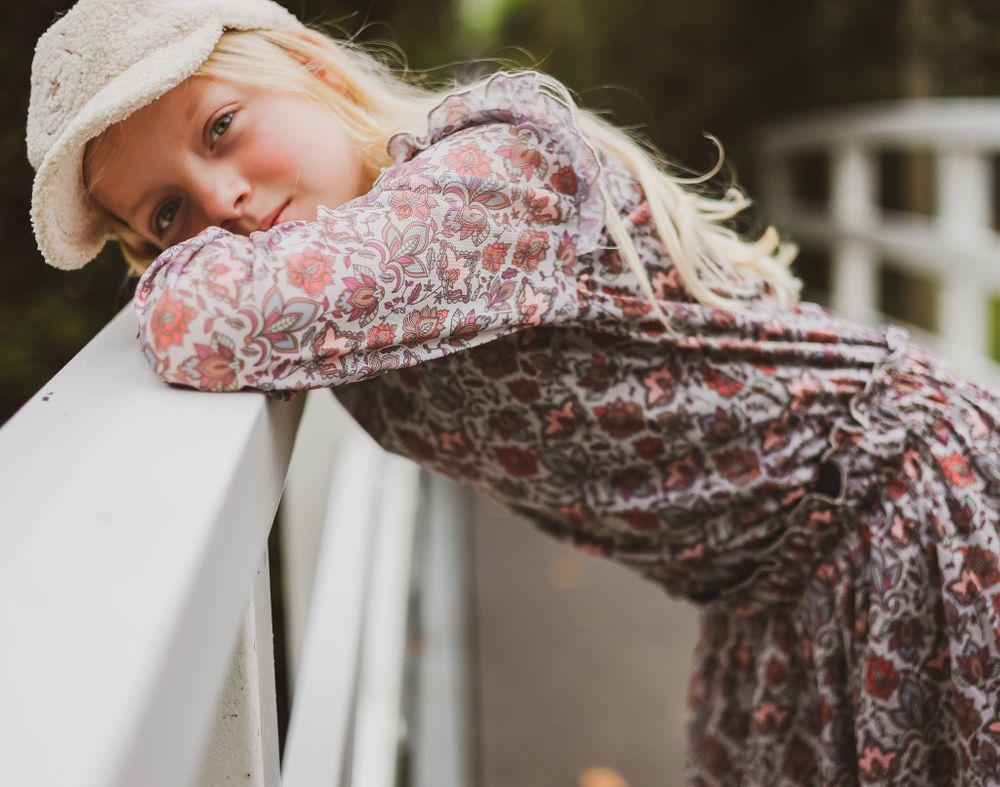 A person in a patterned dress and white cap leans dreamily on a white railing.