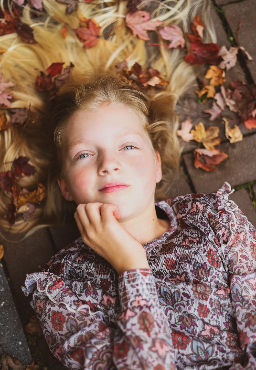 A person in a floral dress lies on ground surrounded by colorful autumn leaves.