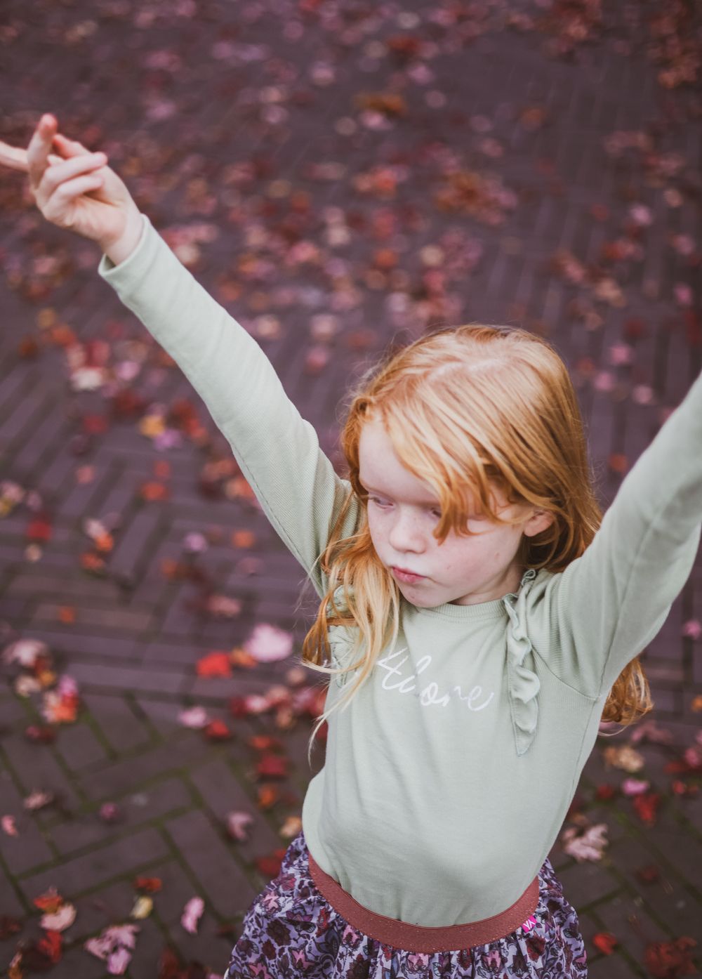A person in a sage green top stretches their arms wide among scattered red autumn leaves.