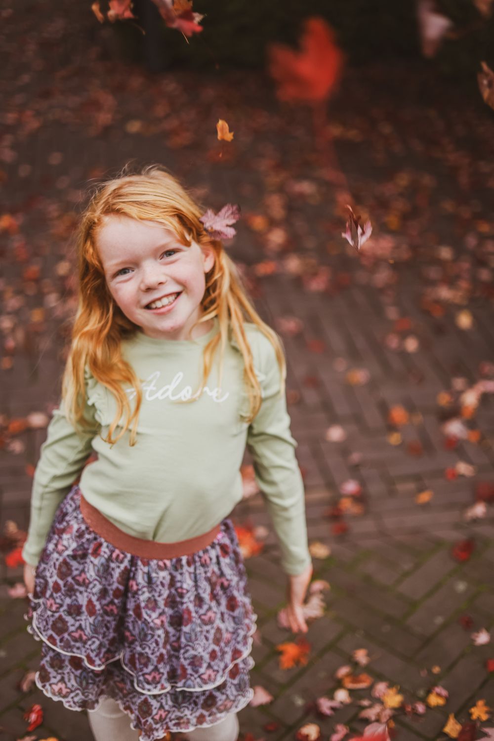 Child in mint sweater and patterned skirt playing with falling autumn leaves.