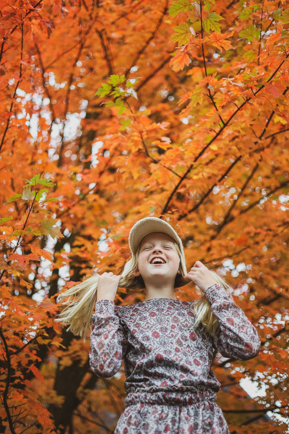 Young person joyfully posing under vibrant orange fall foliage wearing a baseball cap.