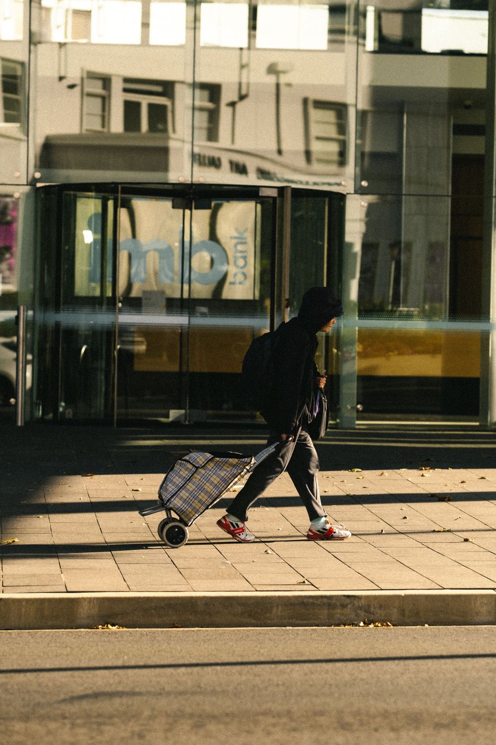Silhouetted figure with shopping cart on tiled sidewalk in urban setting.