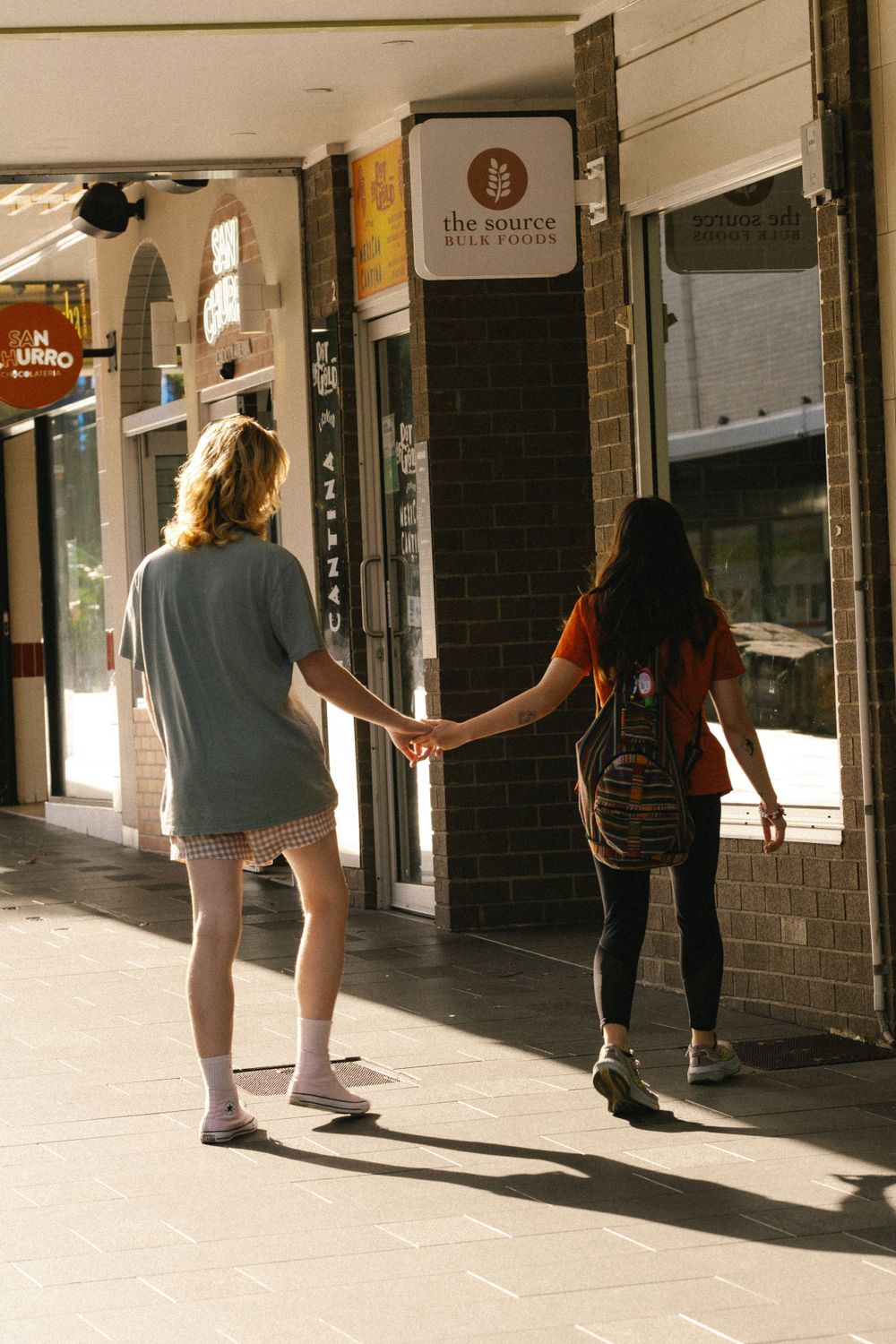 People's legs and feet cast long shadows on a sunlit sidewalk outside storefronts.