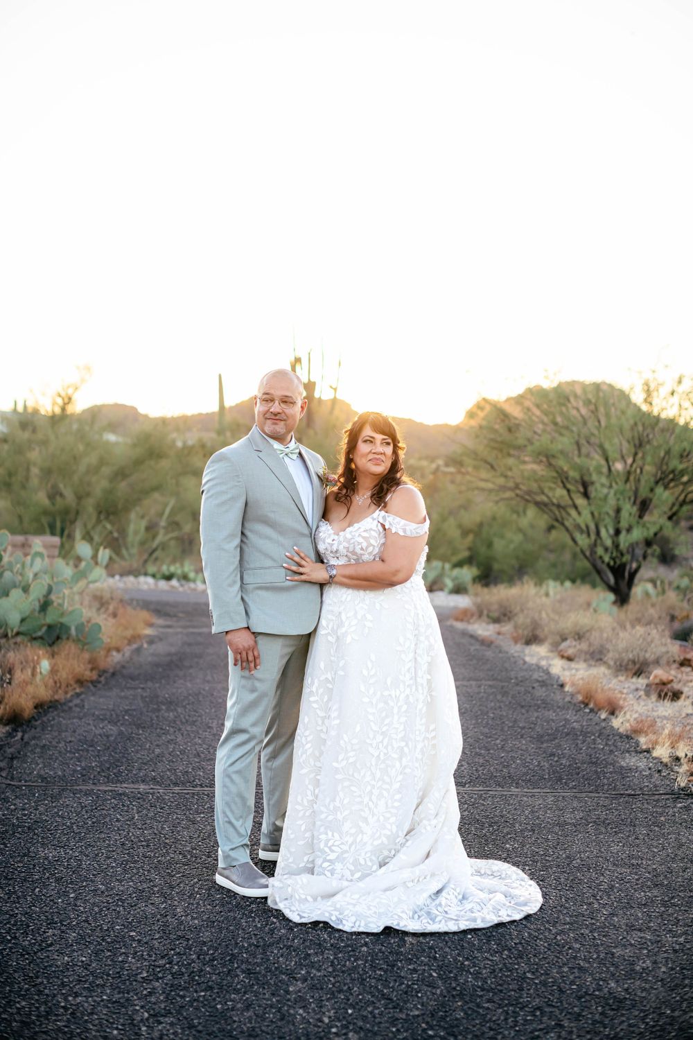 Couple in formal attire posing together on desert path at sunset.