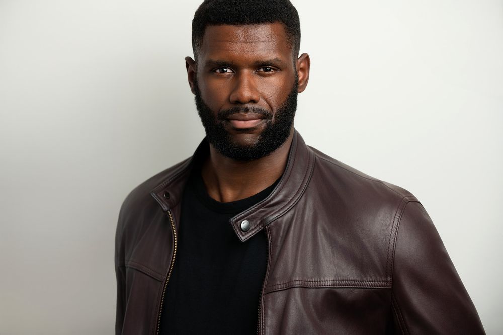 Man in brown leather jacket with beard posing against white background for professional headshot photo.