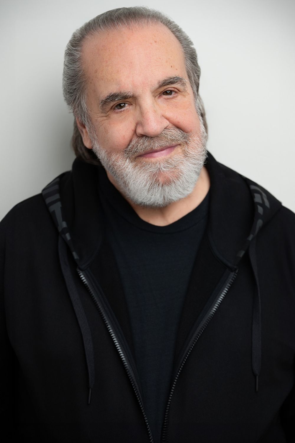 Older man with gray beard and silver hair smiling, wearing a black hoodie, against a white background.