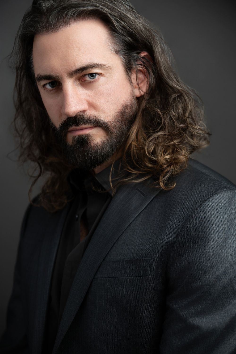 Professional headshot of a bearded man with long curly hair wearing a dark blazer against a gray background.
