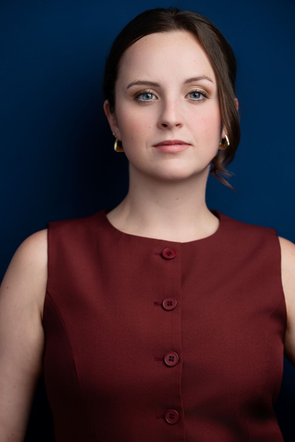 Professional woman in burgundy sleeveless dress with gold hoop earrings poses against dark blue background.