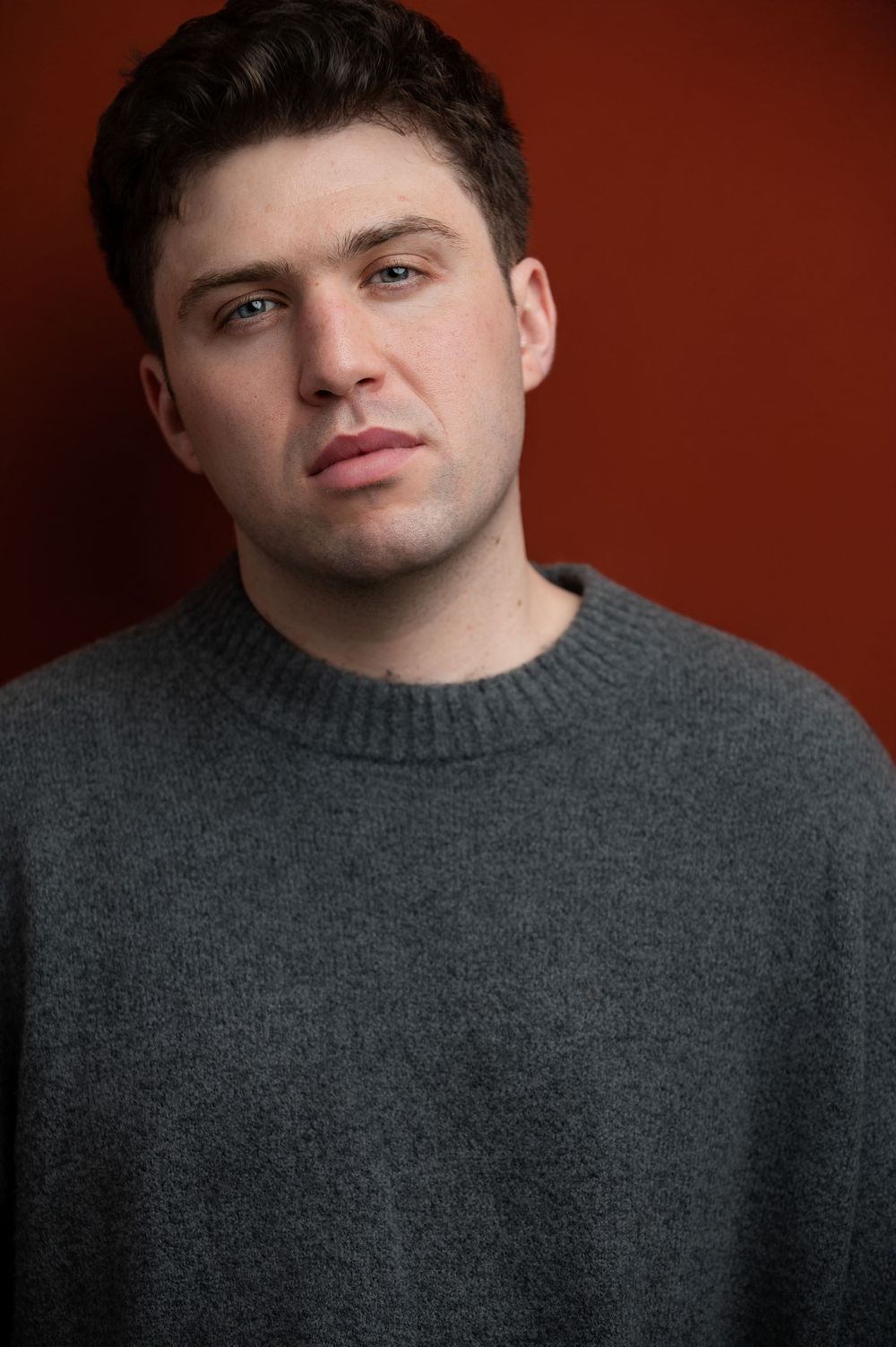 Young man in gray crewneck sweater poses against a red-brown background in a professional headshot photo.