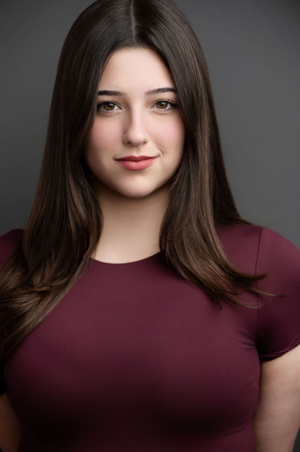 Young woman with long dark hair wearing a burgundy top poses for a professional headshot against a gray background.