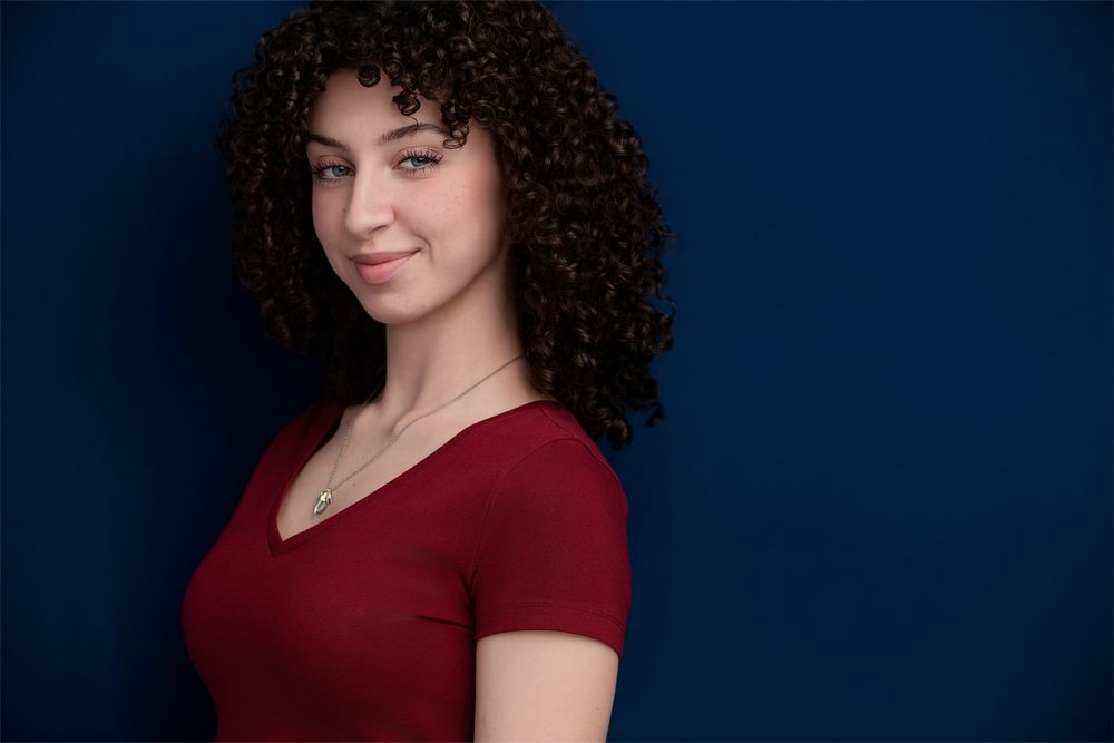Studio actor headshot of a White female actor captured against a clean dark blue backdrop with natural lighting and natural curly hair.