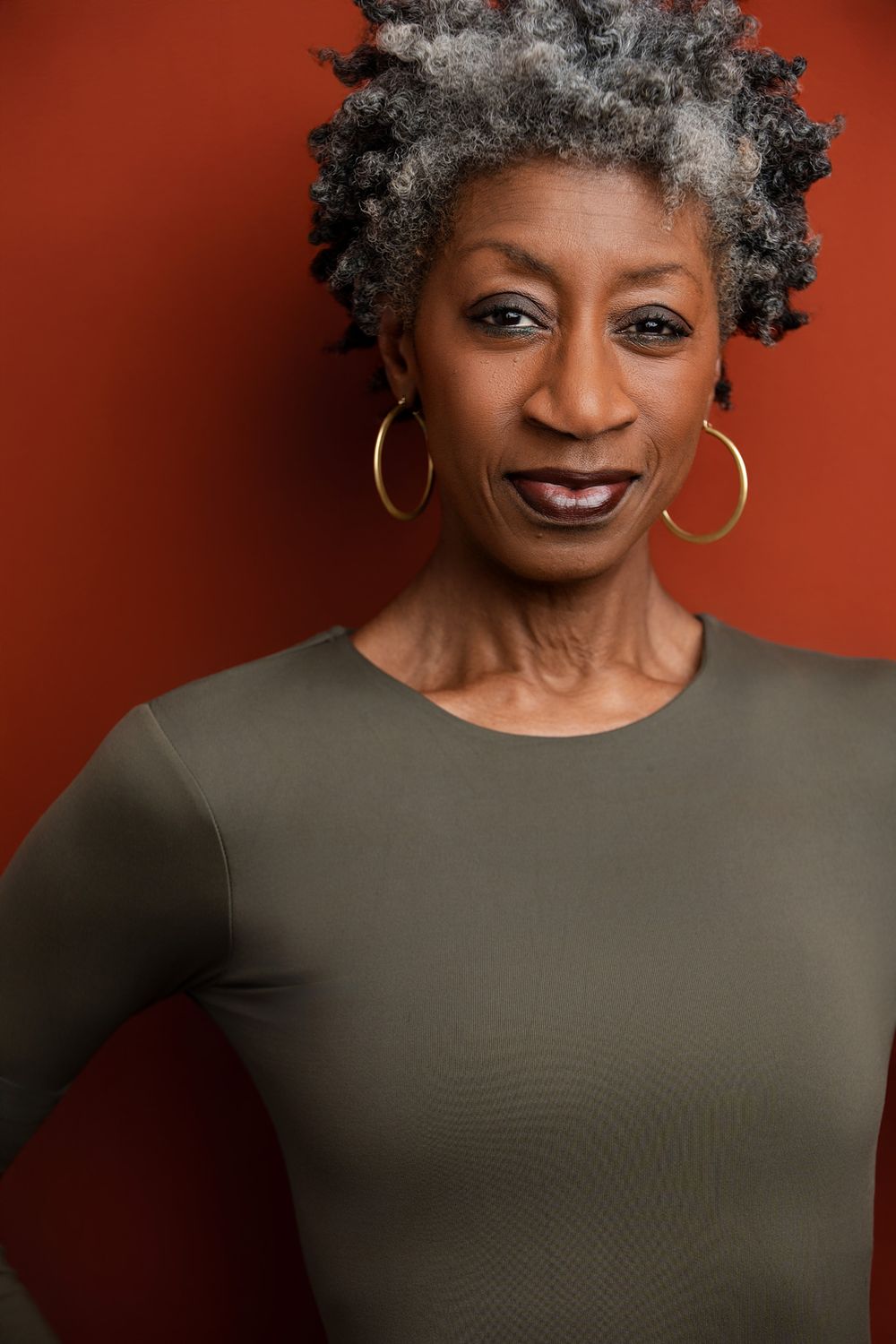 Elegant older Black woman with gray natural hair, gold hoop earrings, and olive top poses against a red-brown background.