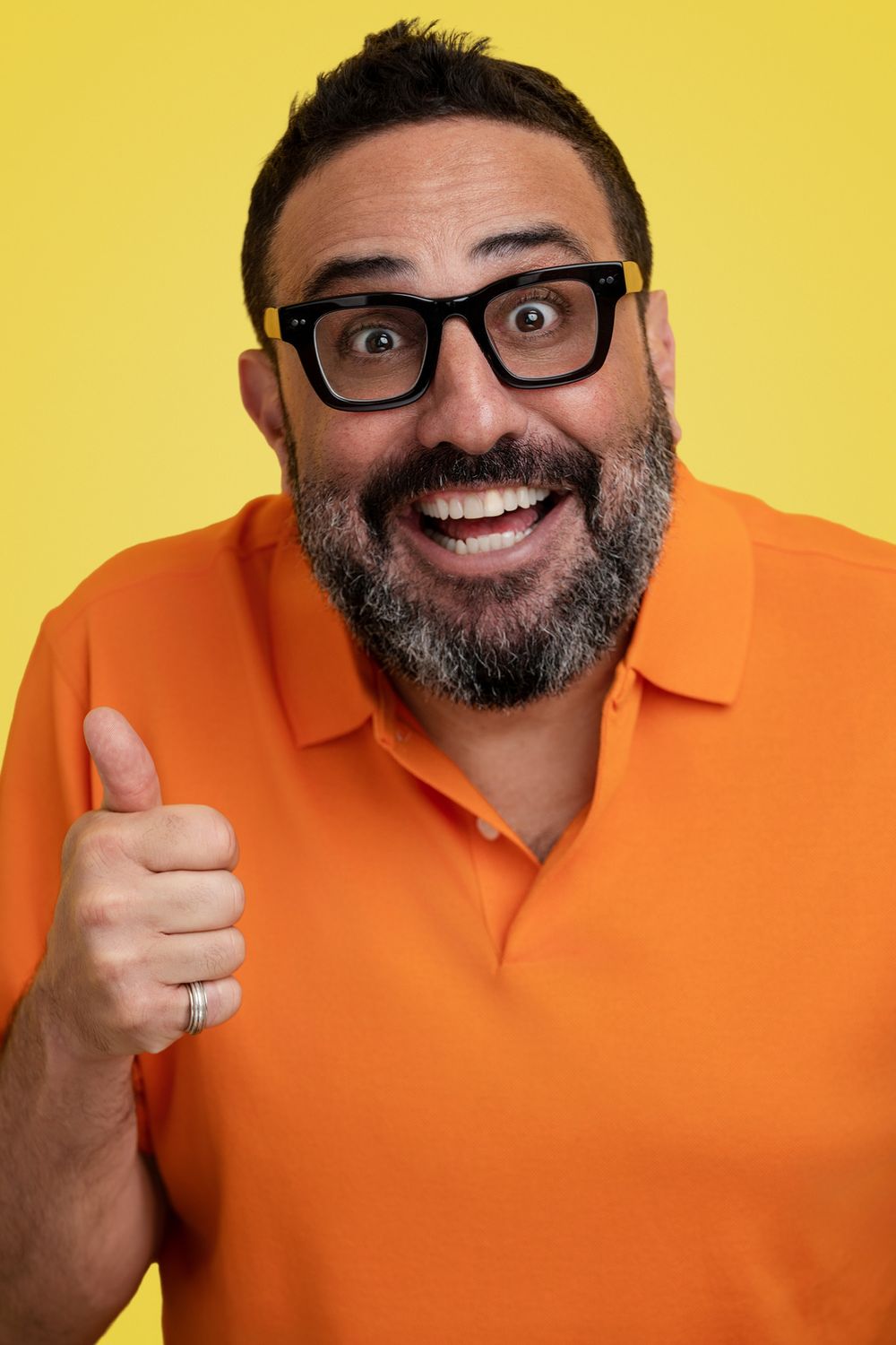 Excited commercial actor wearing glasses and orange polo shirt gives enthusiastic thumbs up against bright yellow background.