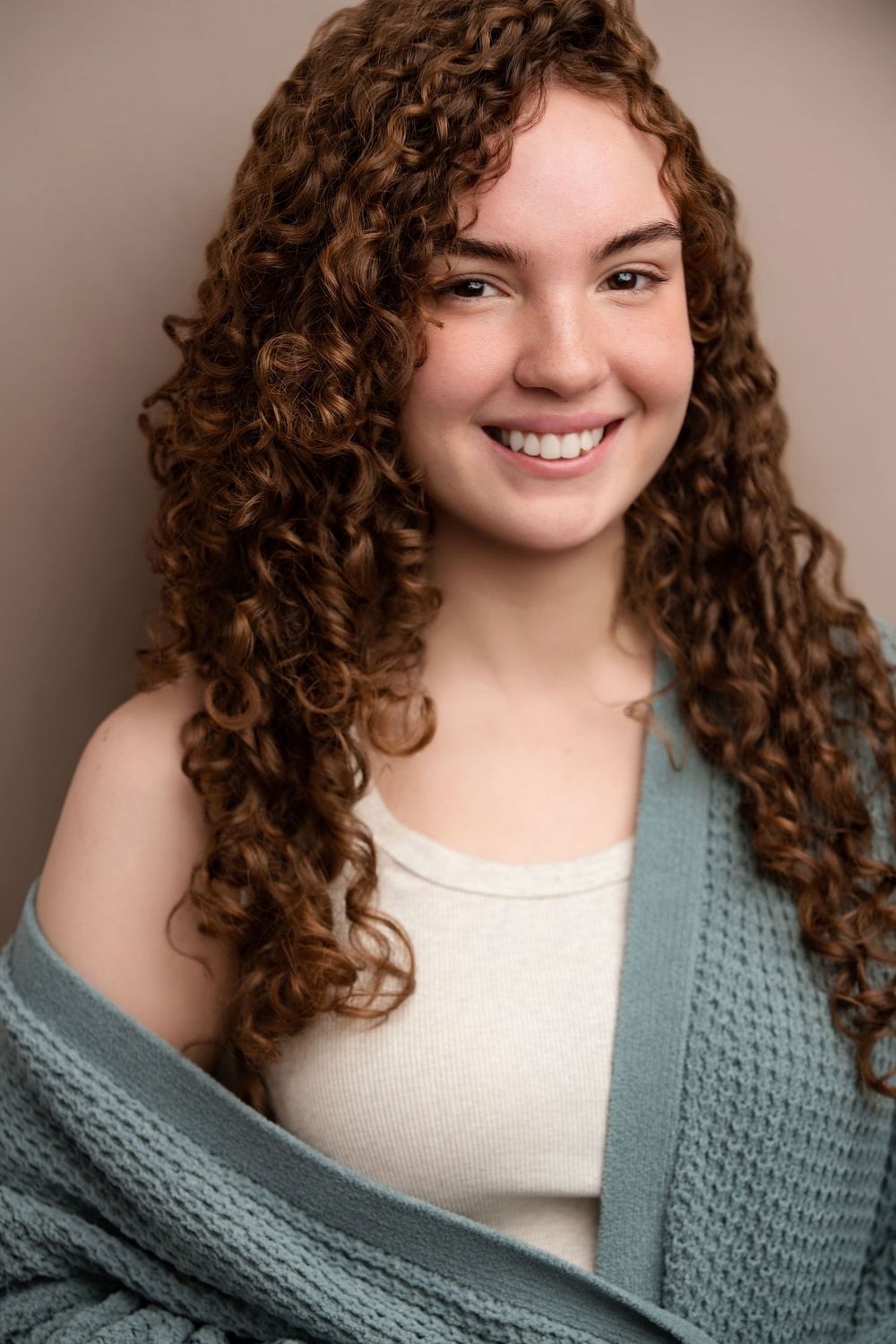 Young woman with curly auburn hair smiling warmly, wearing a sage green cardigan and cream top against a neutral background.