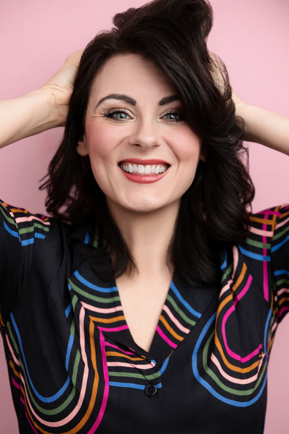 Smiling brunette woman in colorful striped black blouse poses with hands in hair against pink background.