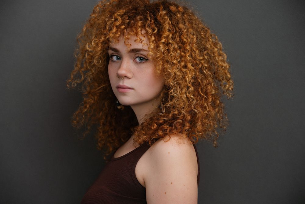 Professional headshot of a mixed race female actor captured on dark background featuring stunning natural curly red hair and brown tank top.