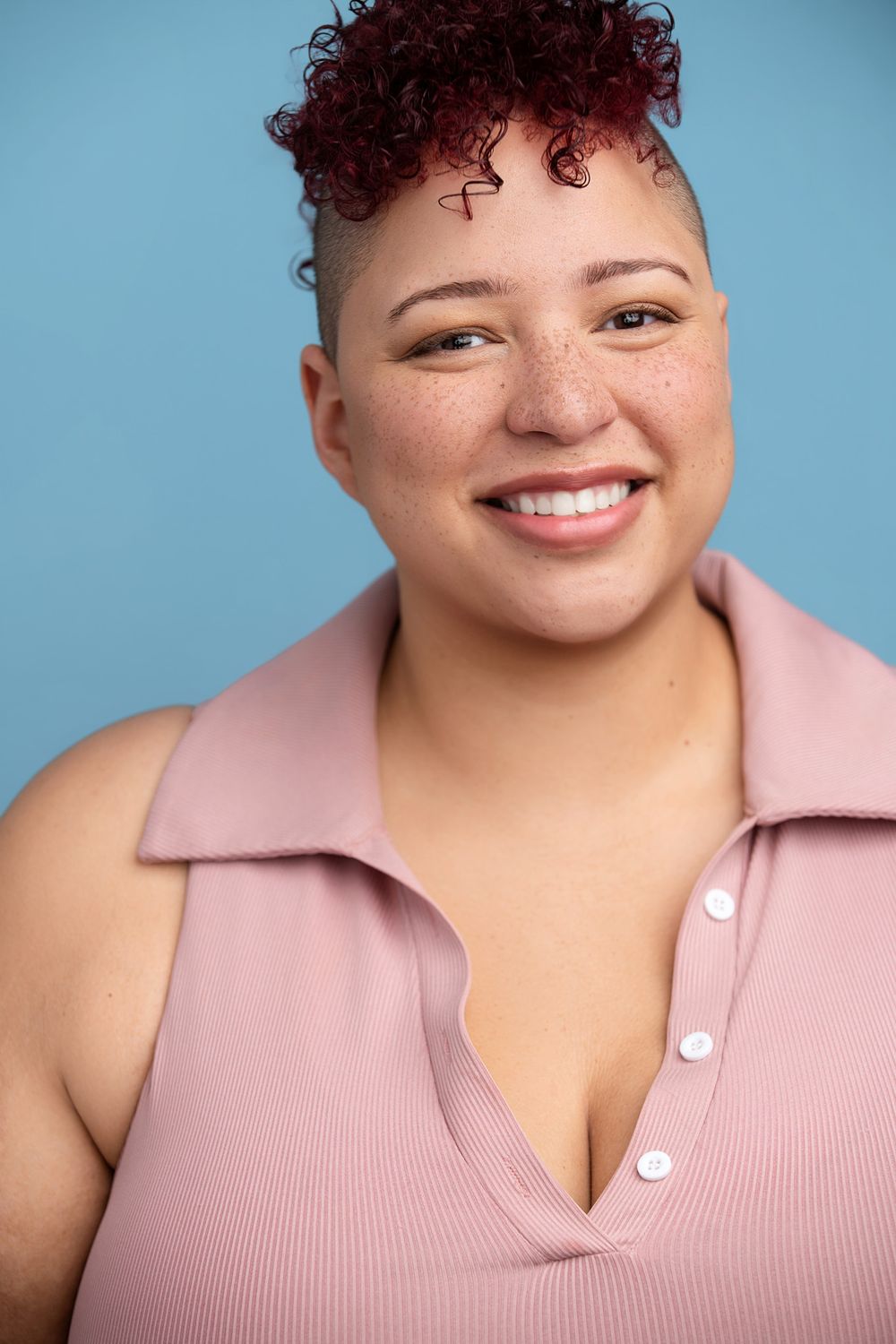 Smiling woman with red curly hair wearing a pink sleeveless top poses against a blue background.