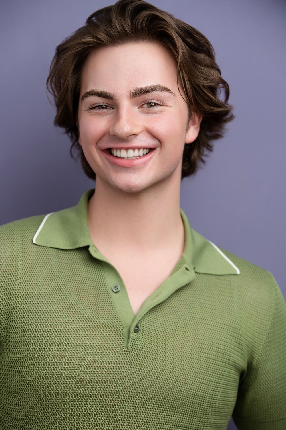 Smiling young man with wavy brown hair wearing a green polo shirt against a purple background.