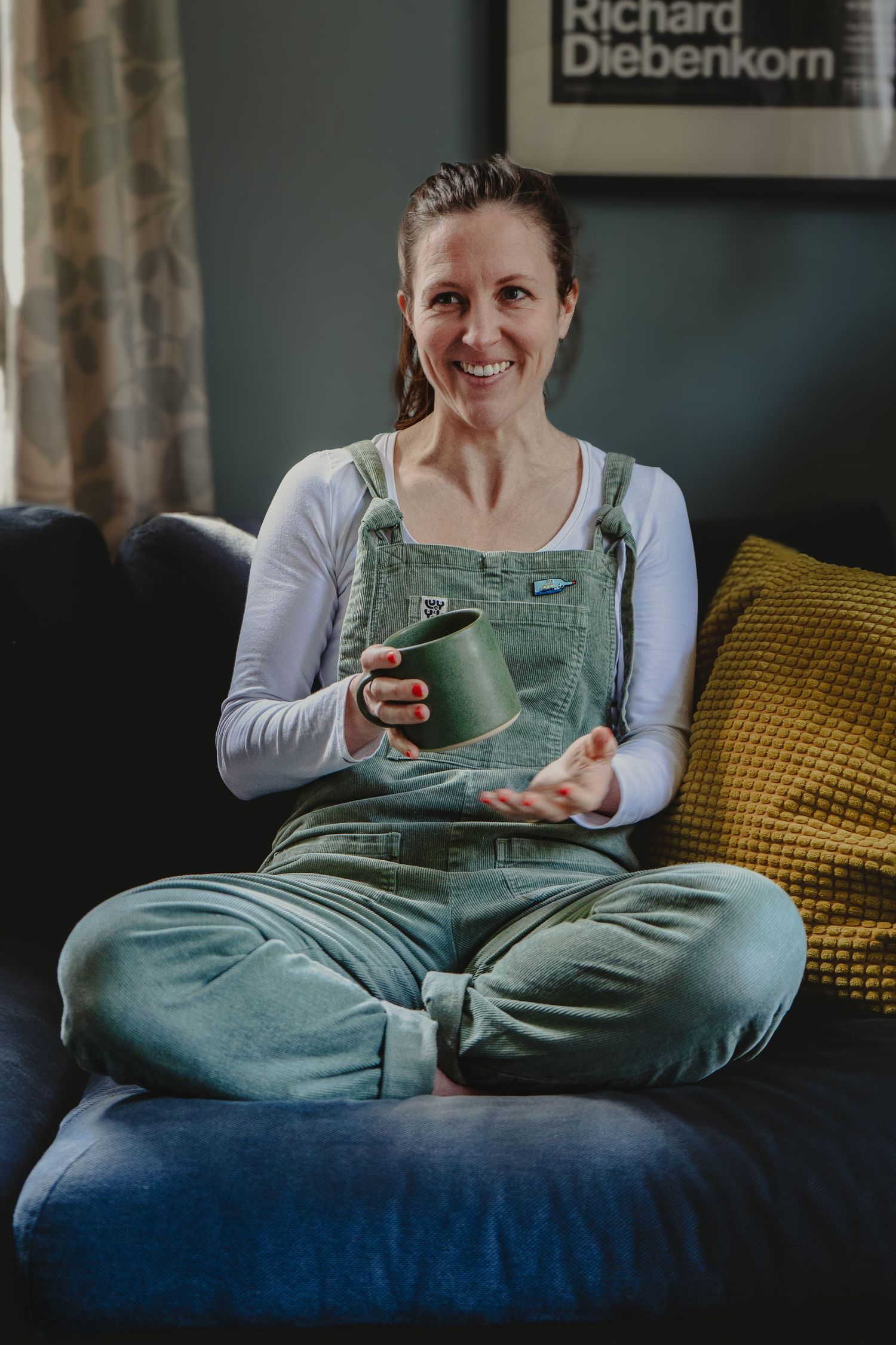 Brand portrait of a female illustrator dressed in green, sat on a blue sofa with a cup in hand, mid conversation and laughing.