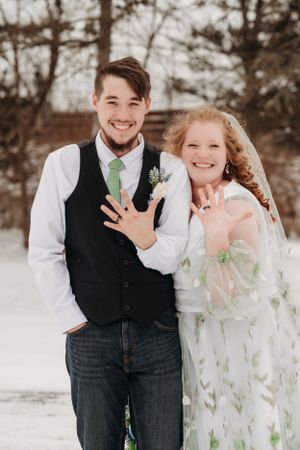Winter wedding portrait of a newly married couple showing off their rings in a snowy outdoor setting.