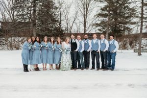 Wedding party poses in light blue attire against snowy winter backdrop with evergreen trees.