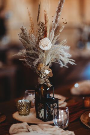 Rustic wedding centerpiece featuring dried flowers and pampas grass in a dark bottle with lantern accents.