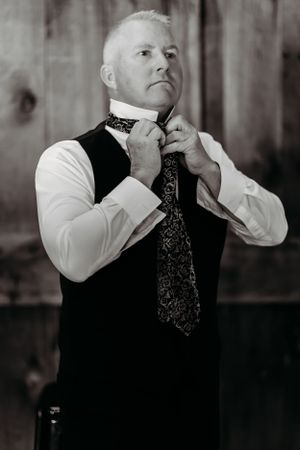 Classic black and white portrait of groom adjusting his patterned tie while wearing a vest.