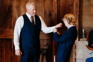 A touching moment between two people in formal attire as one puts on a suit jacket for the other in a rustic wood room.
