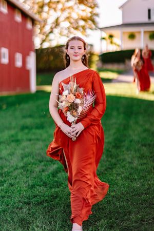 Bridesmaid in flowing orange dress carries dried flower bouquet against rustic farm backdrop.