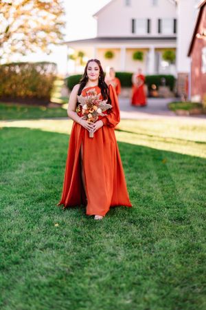 A bridesmaid in a flowing burnt orange dress stands on grass in front of a white farmhouse.